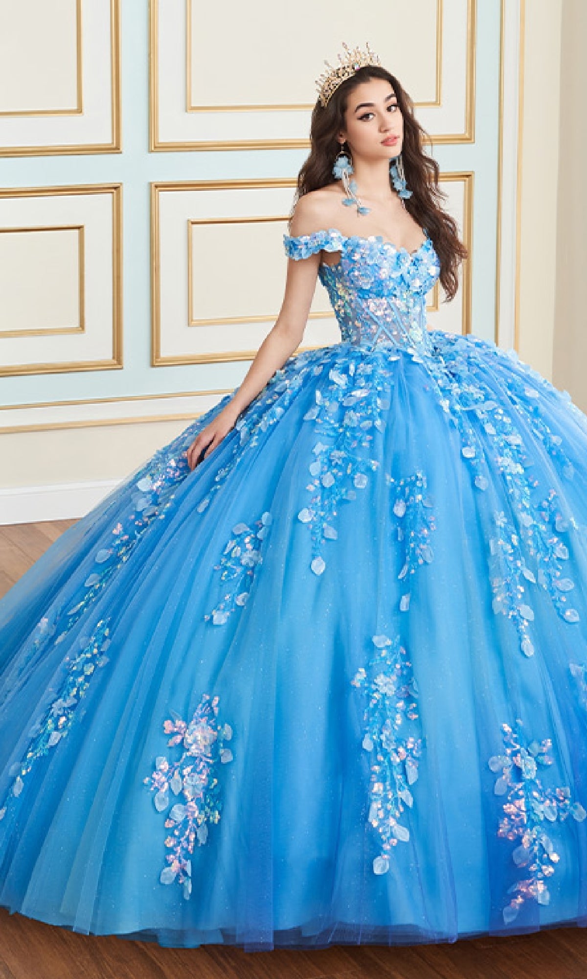 A woman wears the Quinceanera Dress PR30162 By Princesa, a blue off-the-shoulder gown with floral embellishments and glitter tulle accents, standing against a cream and gold paneled wall with a tiara.